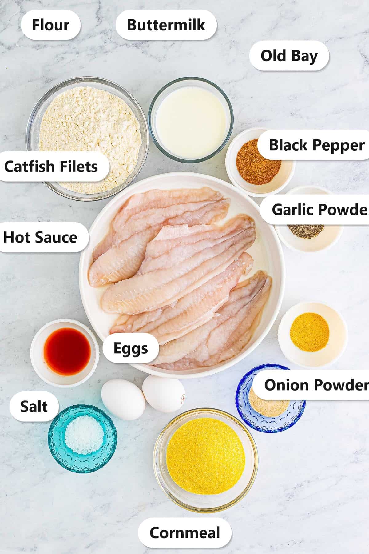 Labeled ingredients flat lay for fried catfish recipe showing catfish filets, flour, buttermilk, Old Bay, black pepper, garlic powder, onion powder, eggs, salt, hot sauce, and cornmeal.