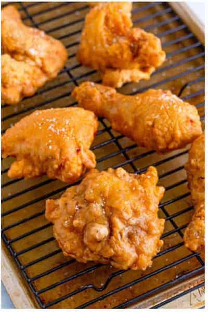 Crispy fried chicken pieces draining on a wire rack over a sheet tray sprinkled with kosher salt.