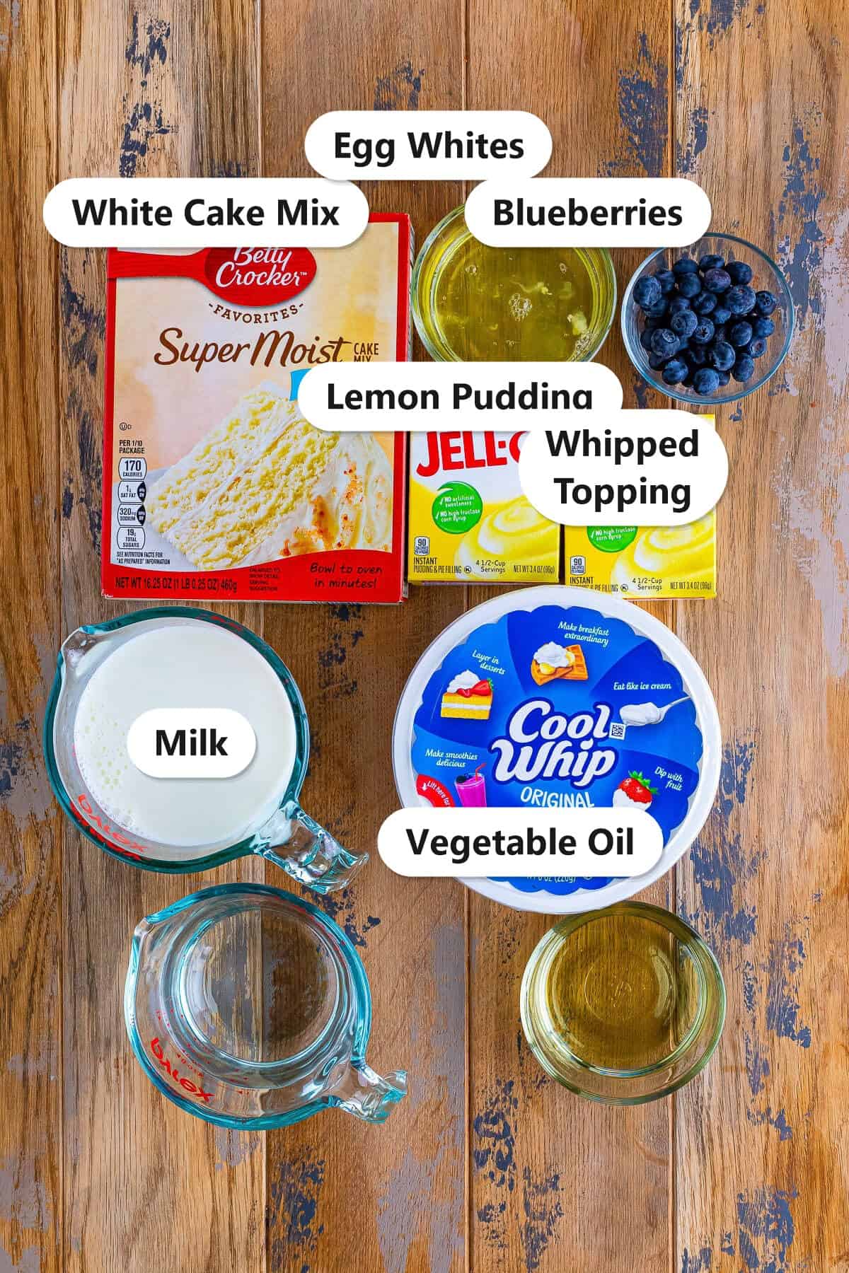Labeled ingredients to make lemon blueberry poke cake including white cake mix, egg whites, blueberries, lemon pudding, whipped topping, milk, and vegetable oil.