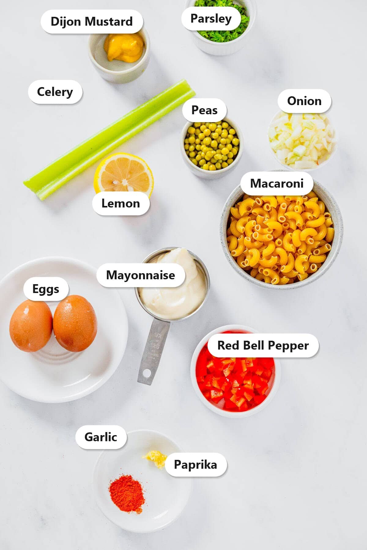 Labeled ingredients for creamy macaroni salad including macaroni peas red bell pepper onion celery eggs mayonnaise lemon dijon and paprika.