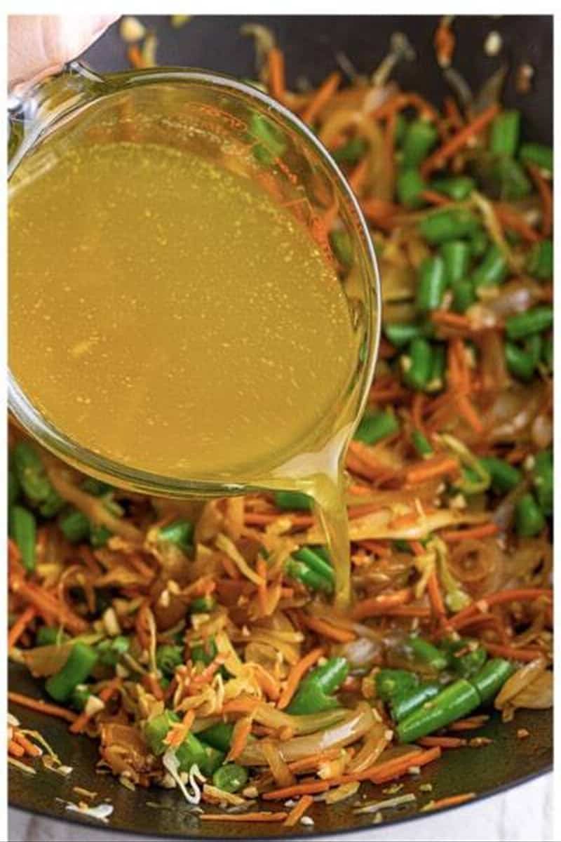Chicken stock being poured into a wok with cooked vegetables for pancit recipe.