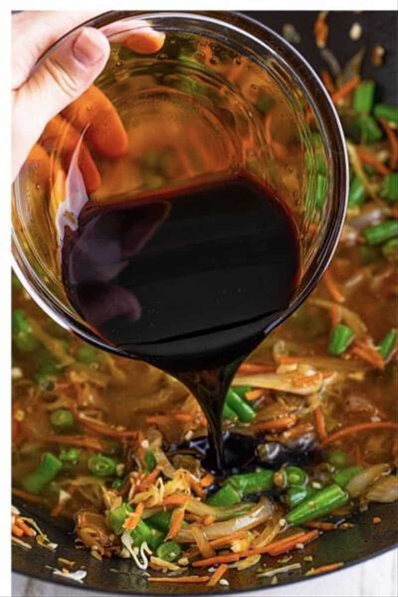 Dark soy sauce mixture being poured into a wok with sauteed pancit vegetables for pancit recipe.