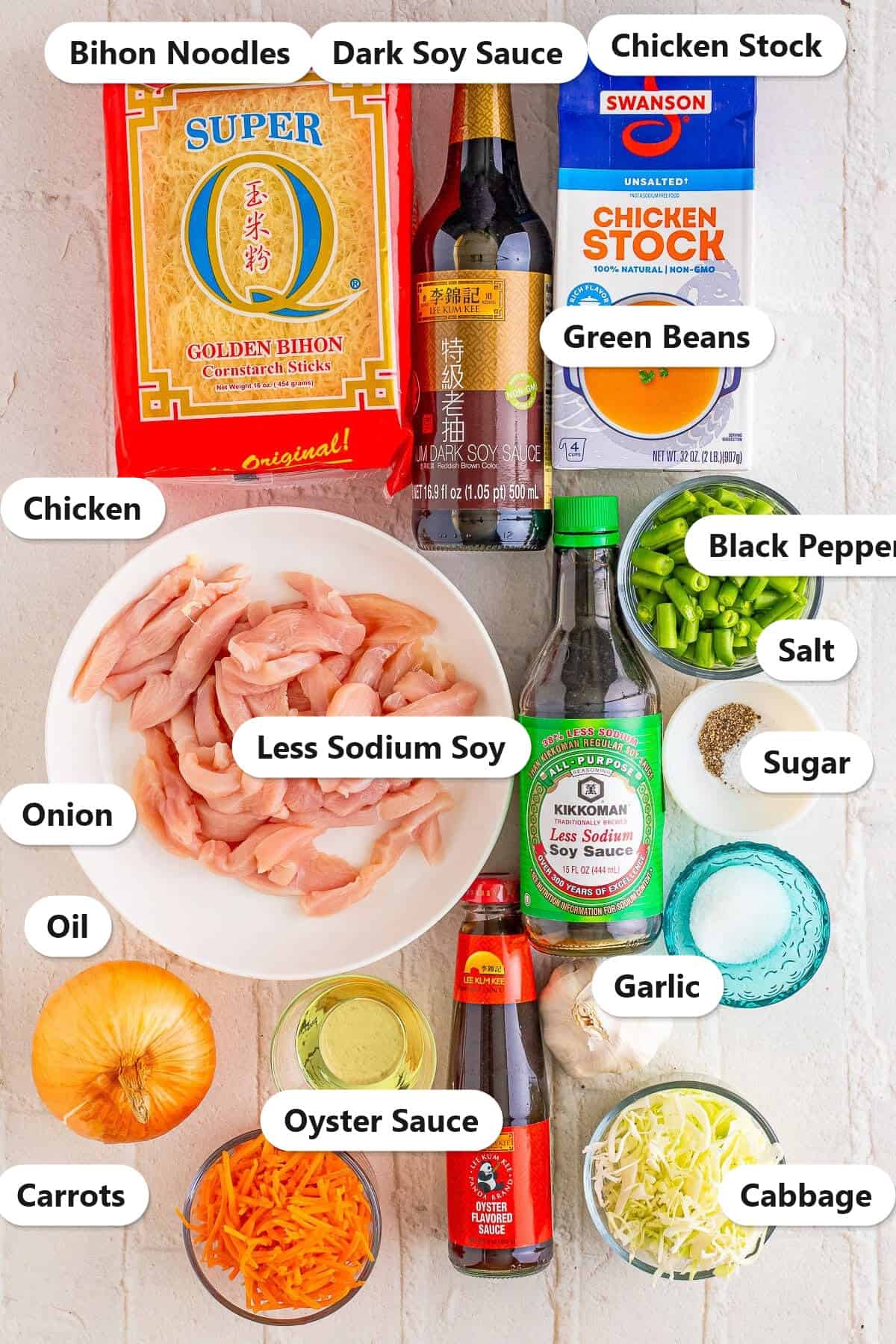 Labeled ingredients flat lay for pancit recipe showing bihon noodles, dark soy sauce, chicken stock, chicken, less sodium soy sauce, green beans, black pepper, salt, sugar, onion, oil, oyster sauce, garlic, carrots, and cabbage.