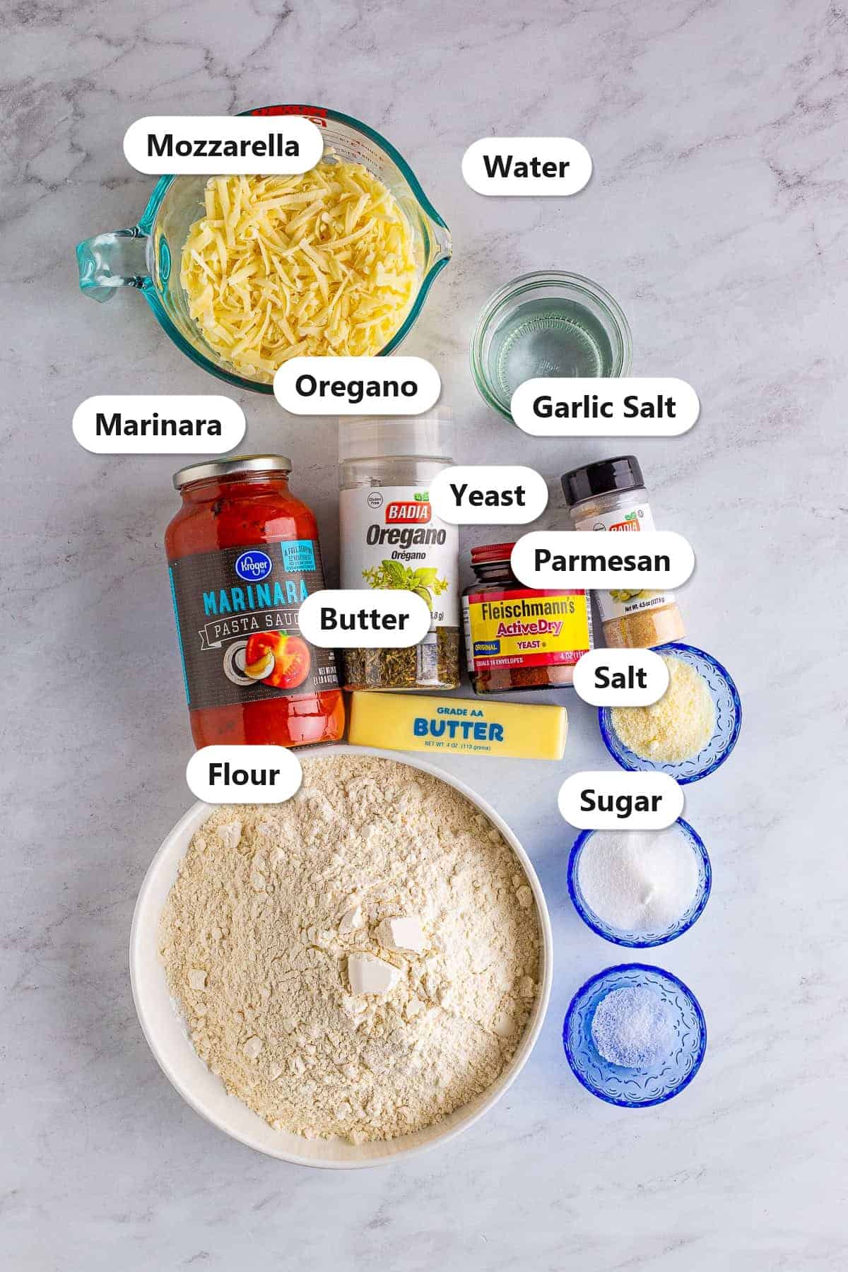 Labeled ingredients to make Pizza Hut cheese sticks including mozzarella, parmesan, flour, yeast, butter, sugar, garlic salt, oregano, salt, water, and marinara.
