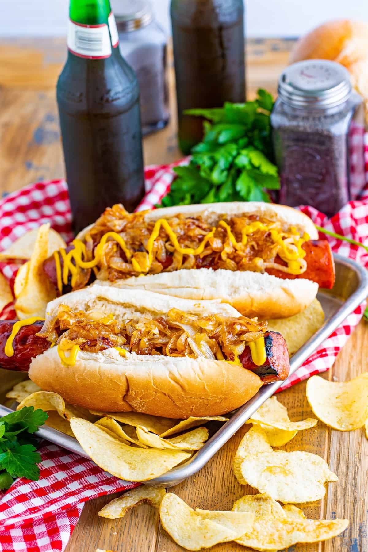 Two finished Polish Sausage Sandwich Recipe sandwiches plated with potato chips on a red checkered cloth.