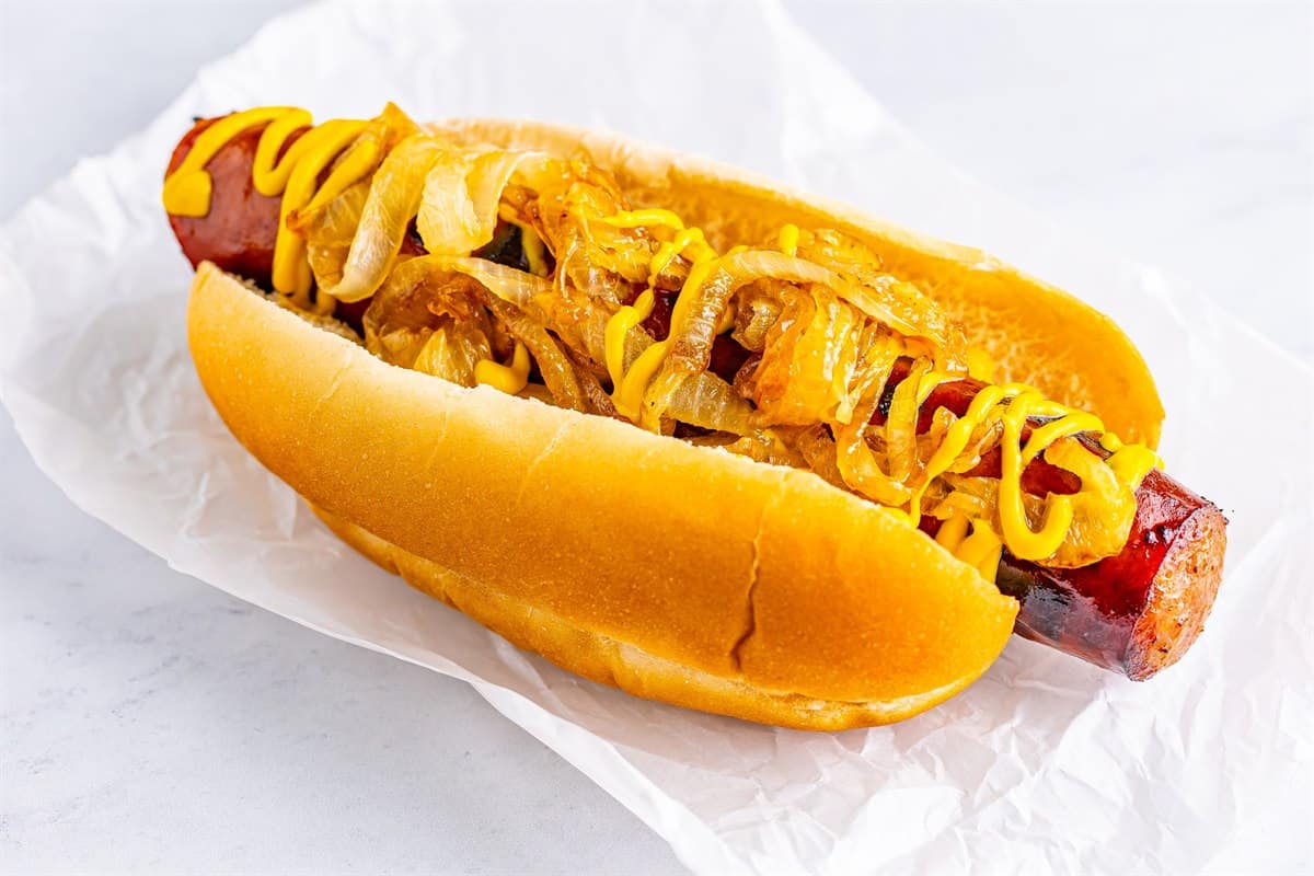 An assembled Polish Sausage Sandwich Recipe with mustard and grilled onions on a toasted hoagie roll wrapped in paper.