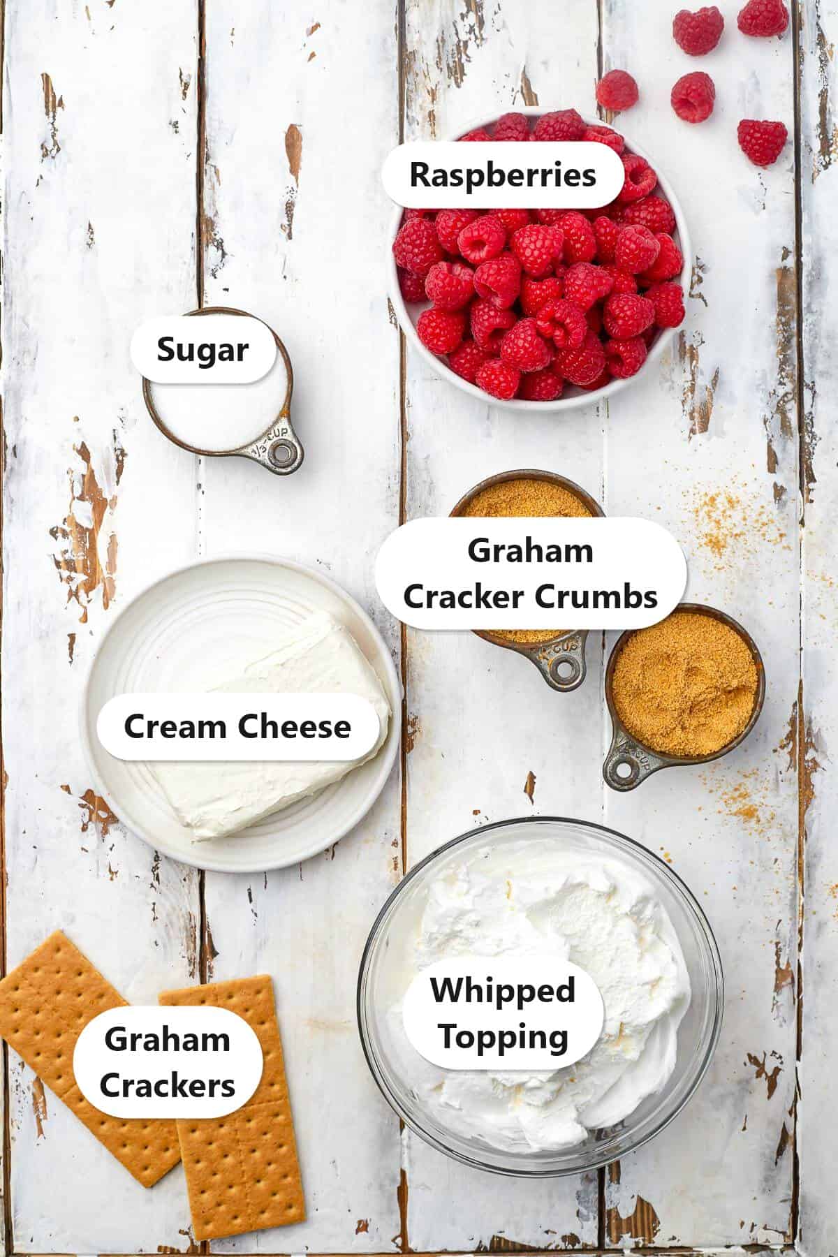 Labeled ingredients to make raspberry cheesecake bites including cream cheese, sugar, fresh raspberries, whipped topping, and graham cracker crumbs.