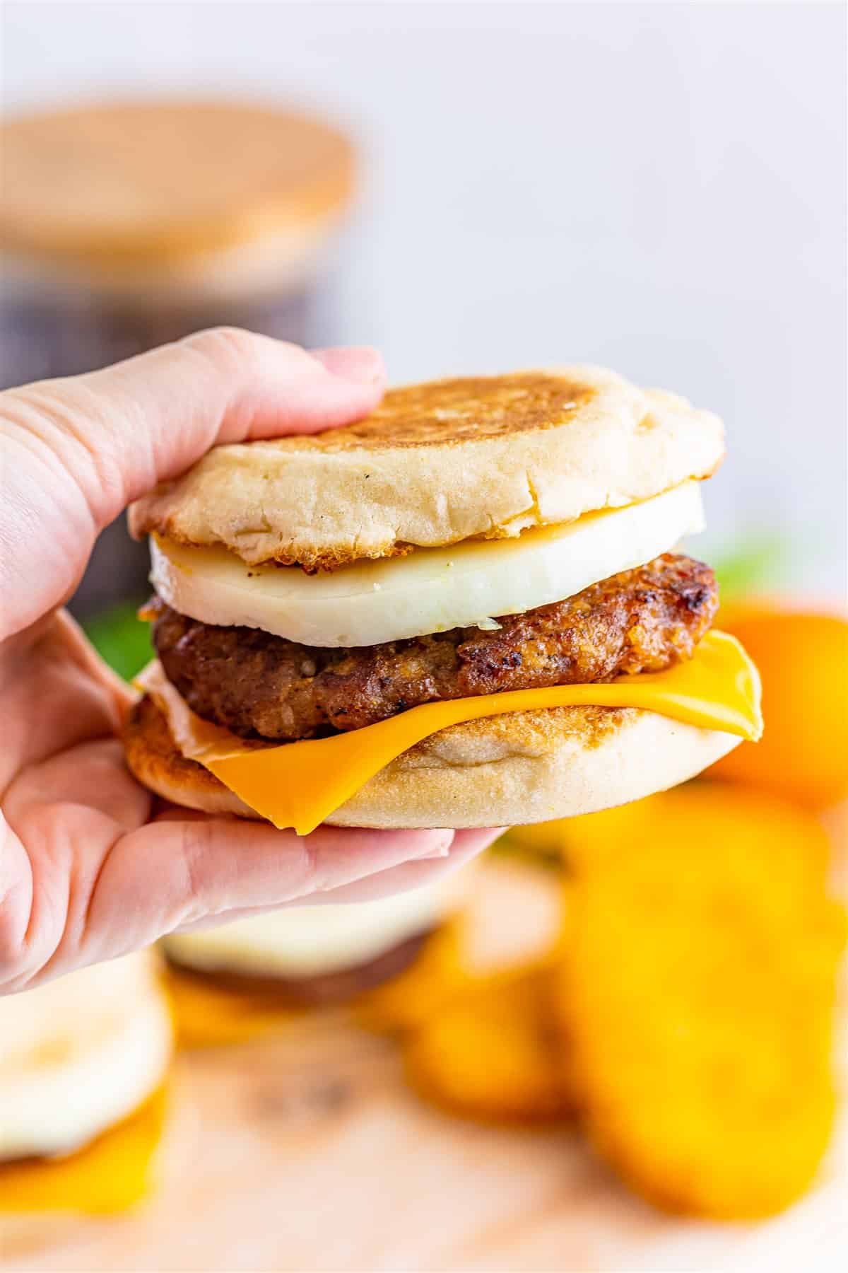 Hand holding a Sausage Egg McMuffin Recipe sandwich showing the layers of cheese, sausage, and round egg with hash browns in the background.