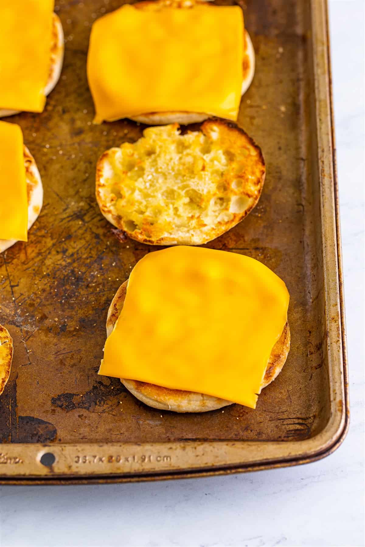 Toasted English muffin halves topped with American cheese slices on a sheet pan for the Sausage Egg McMuffin Recipe.