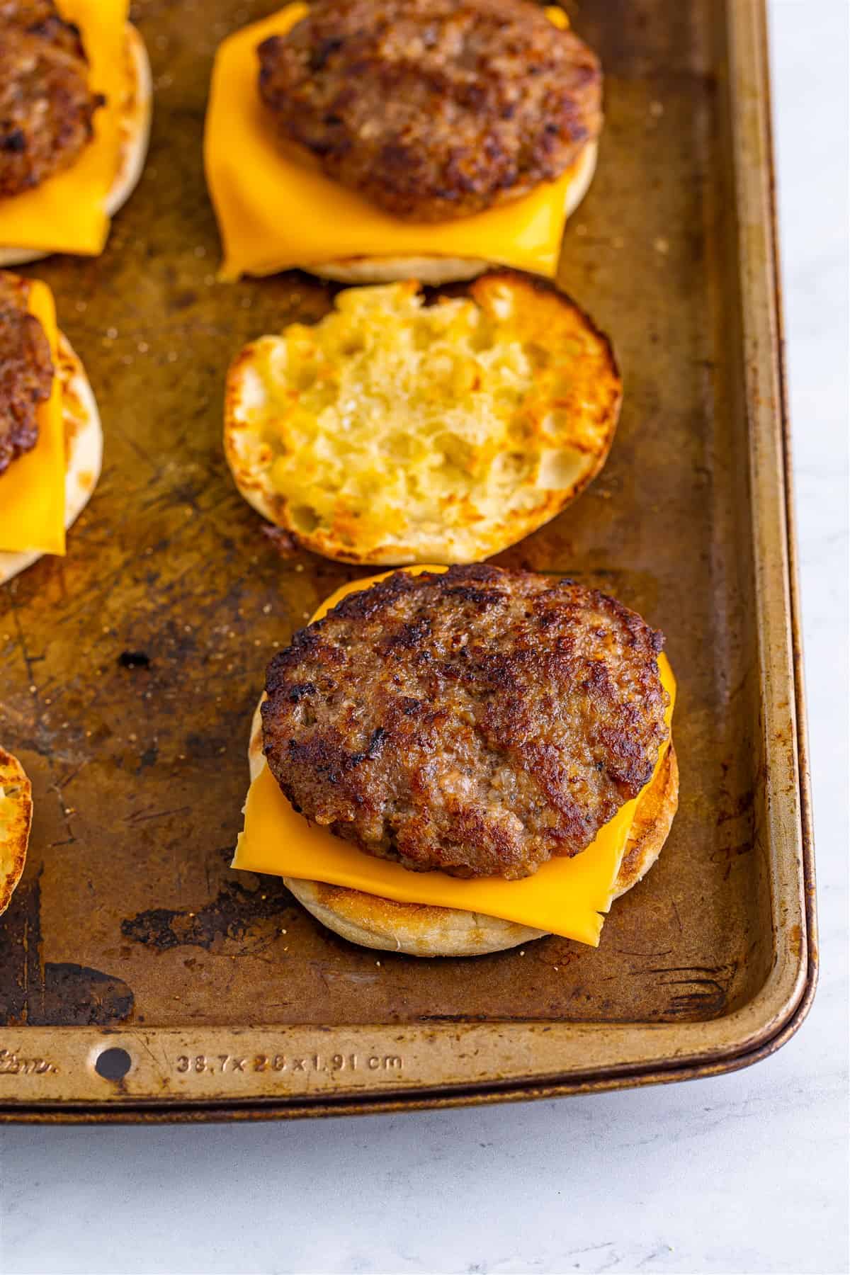 Assembled Sausage Egg McMuffin Recipe sandwich showing sausage, cheese, and toasted English muffin layers on a baking sheet.