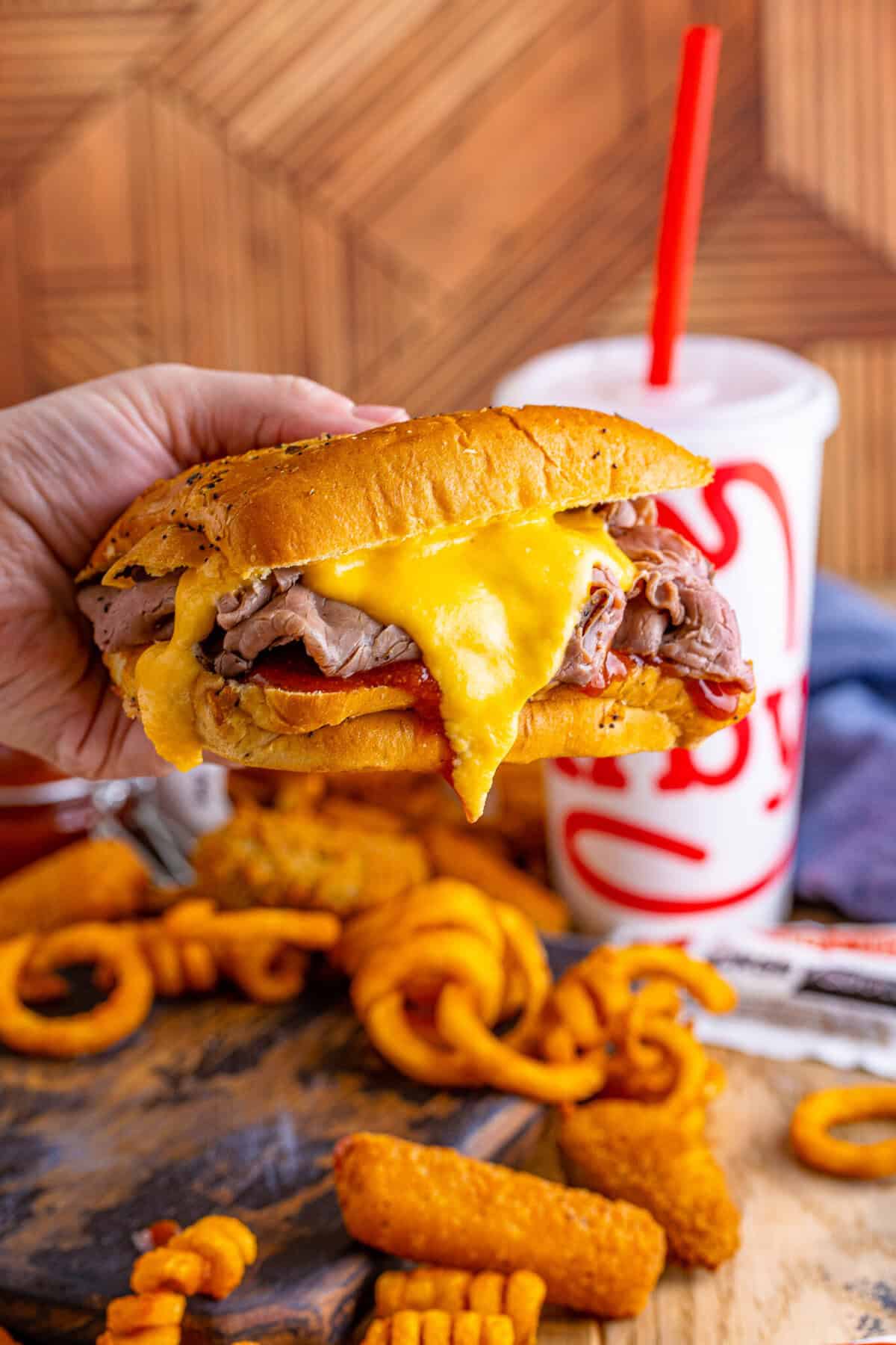 Hand holding a copycat Arbys beef and cheddar sandwich showing the cheese pull and red sauce drip with a fountain drink behind.