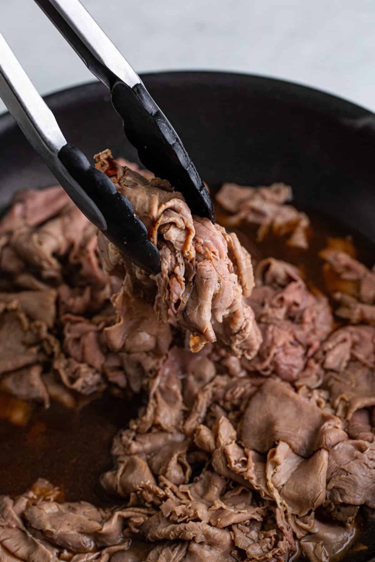 Shaved roast beef warmed in a simmering beef broth in a cast iron skillet being lifted with tongs.