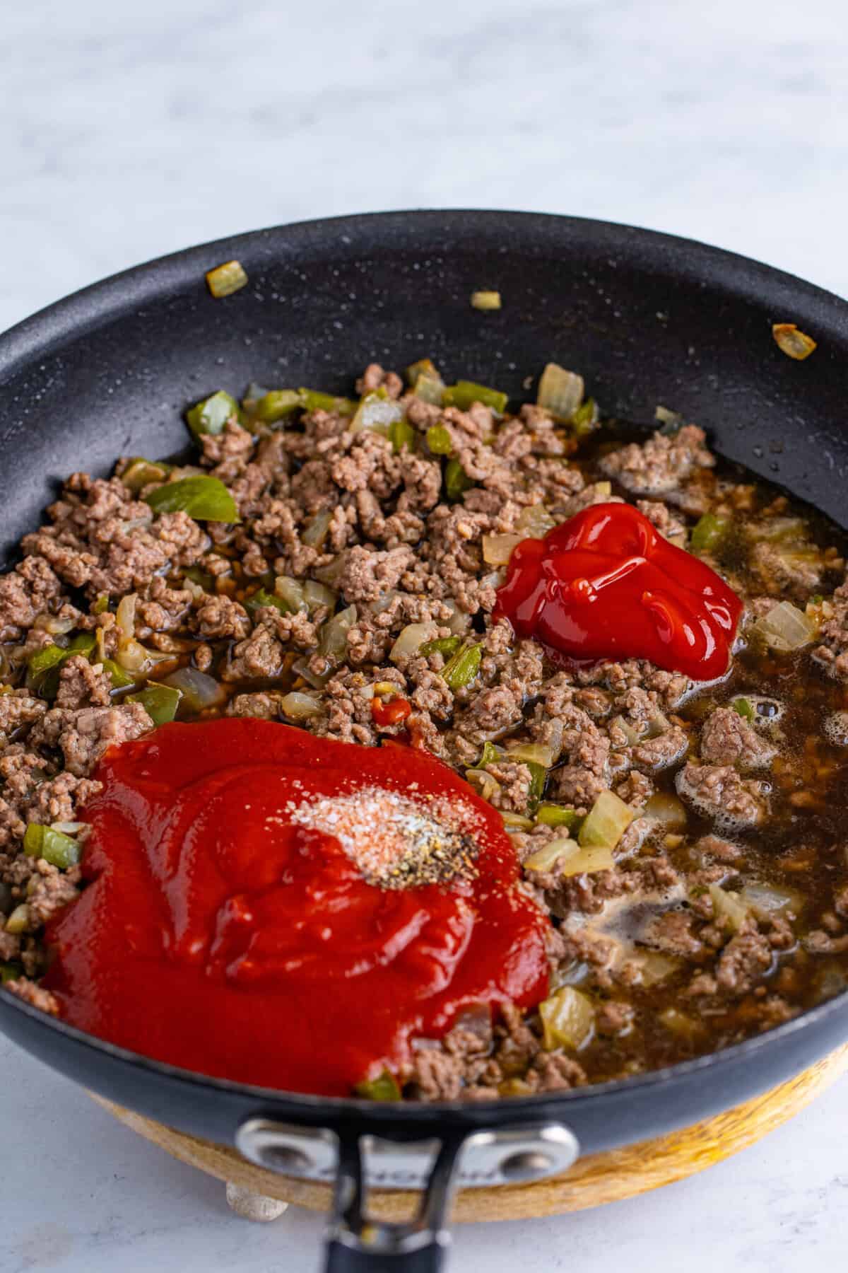 Tomato sauce, ketchup, and Dr Pepper poured over browned ground beef with vegetables in a skillet before stirring and simmering together.