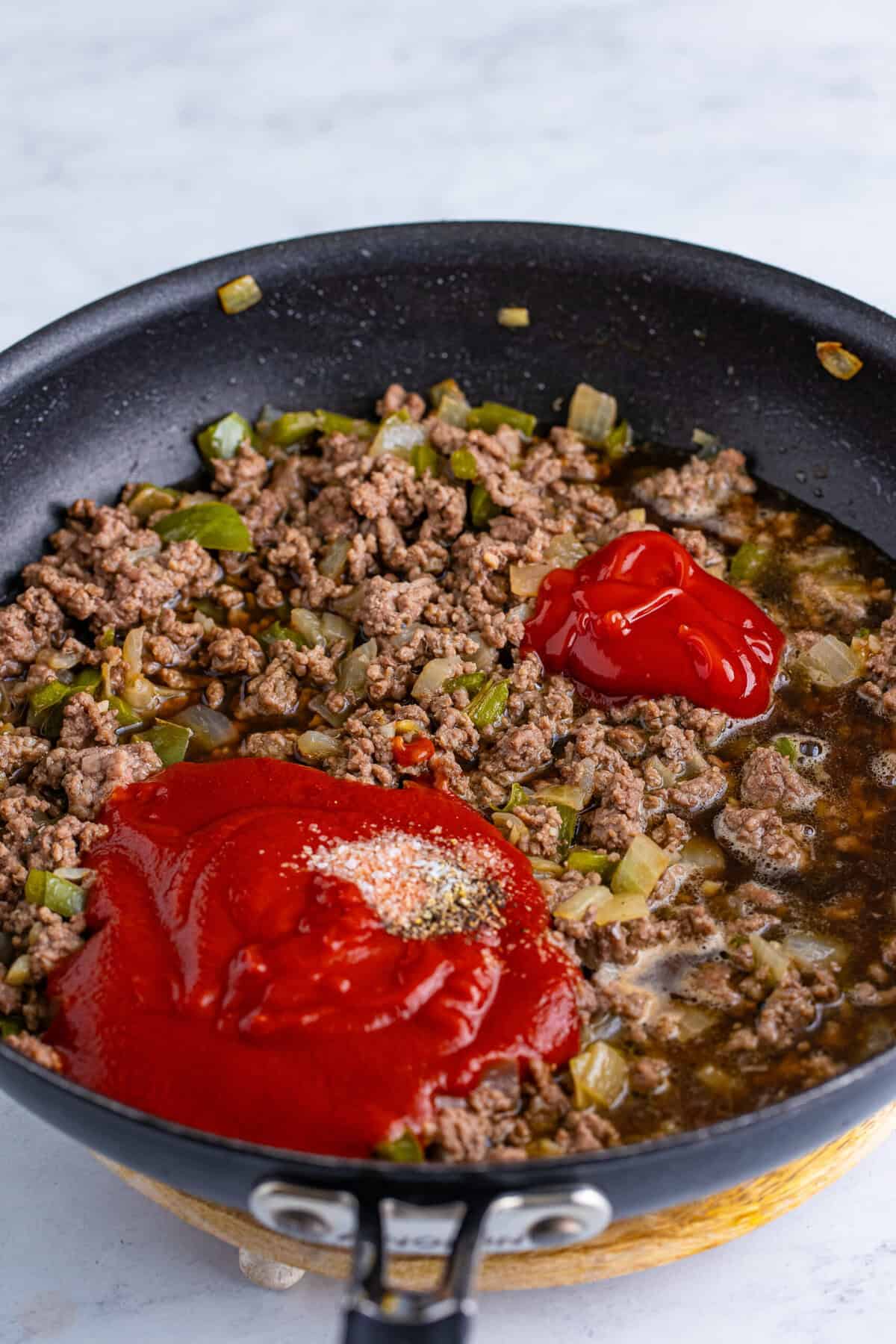 Tomato sauce, ketchup, and Dr Pepper poured over browned ground beef with vegetables in a skillet before stirring and simmering together.