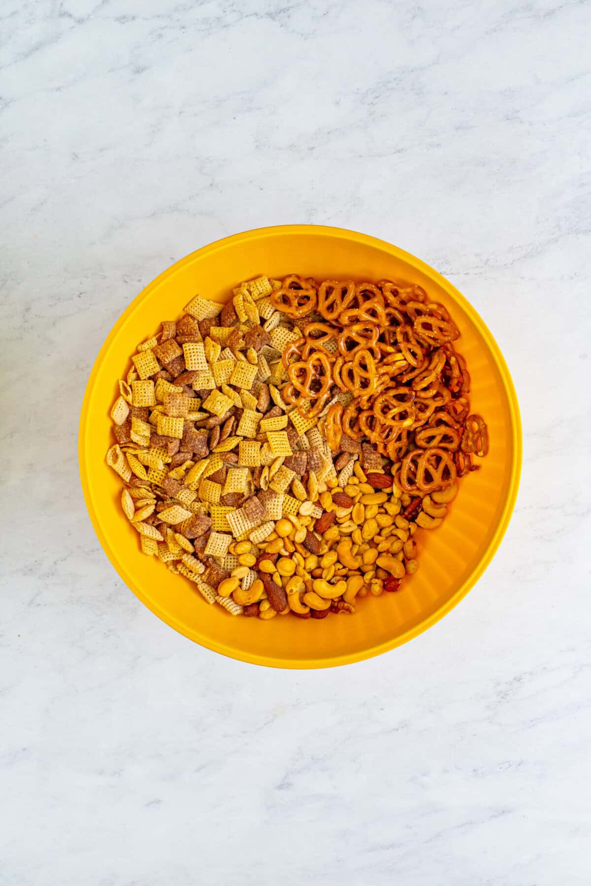 Yellow bowl filled with Chex cereal, mini pretzels, and mixed nuts combined for the Best Sweet Chex Mix Recipe Muddy Buddy.