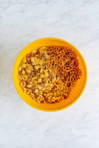 Yellow bowl filled with Chex cereal, mini pretzels, and mixed nuts combined for the Best Sweet Chex Mix Recipe Muddy Buddy.