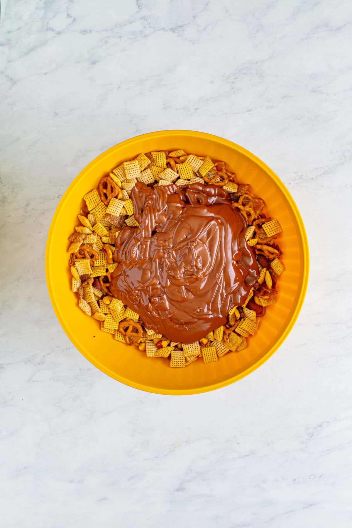 Yellow bowl with chocolate-peanut-butter coating poured over the Chex cereal and pretzel mix before tossing for the Sweet Chex Mix Recipe.