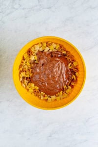 Yellow bowl with chocolate-peanut-butter coating poured over the Chex cereal and pretzel mix before tossing for the Sweet Chex Mix Recipe.