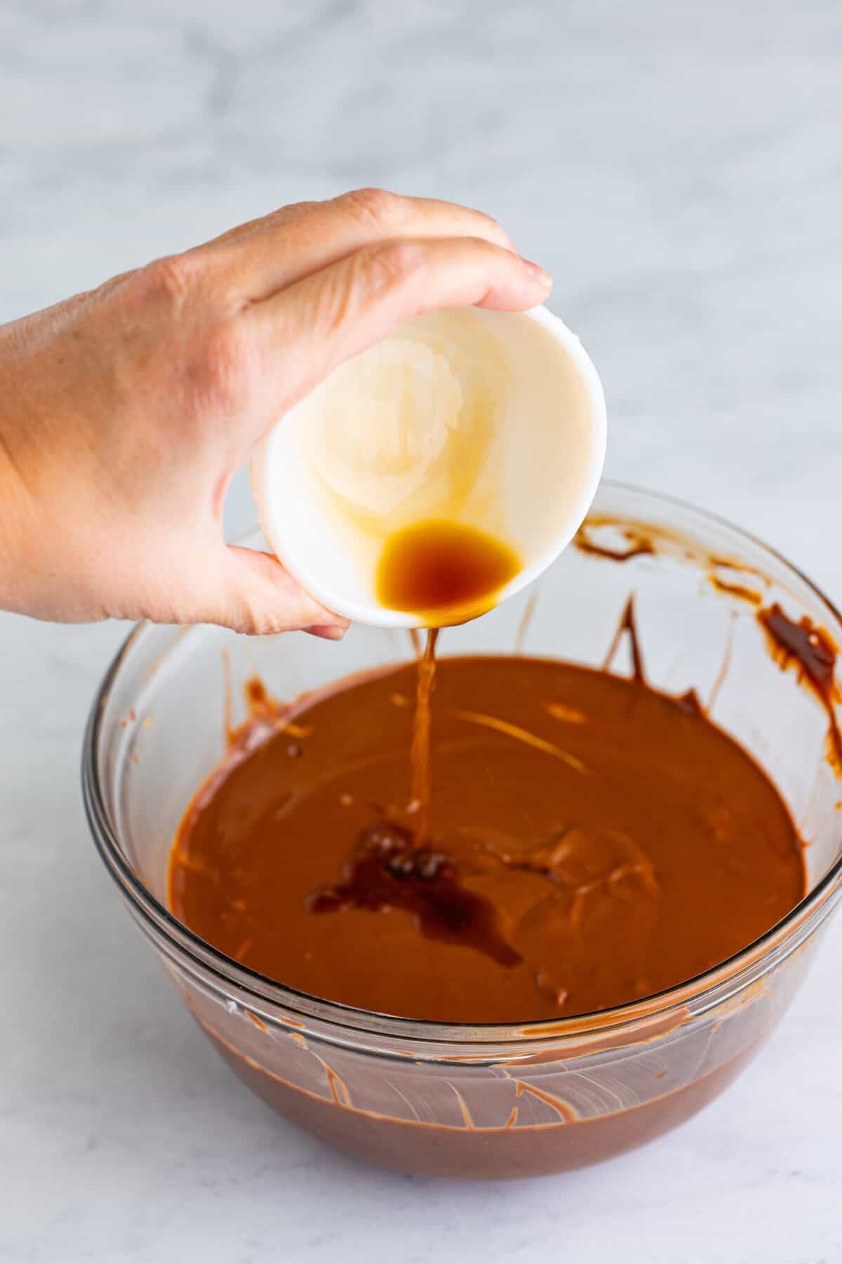 Hand pouring vanilla extract into a glass bowl of smooth melted chocolate-peanut-butter coating for the Sweet Chex Mix Muddy Buddy.