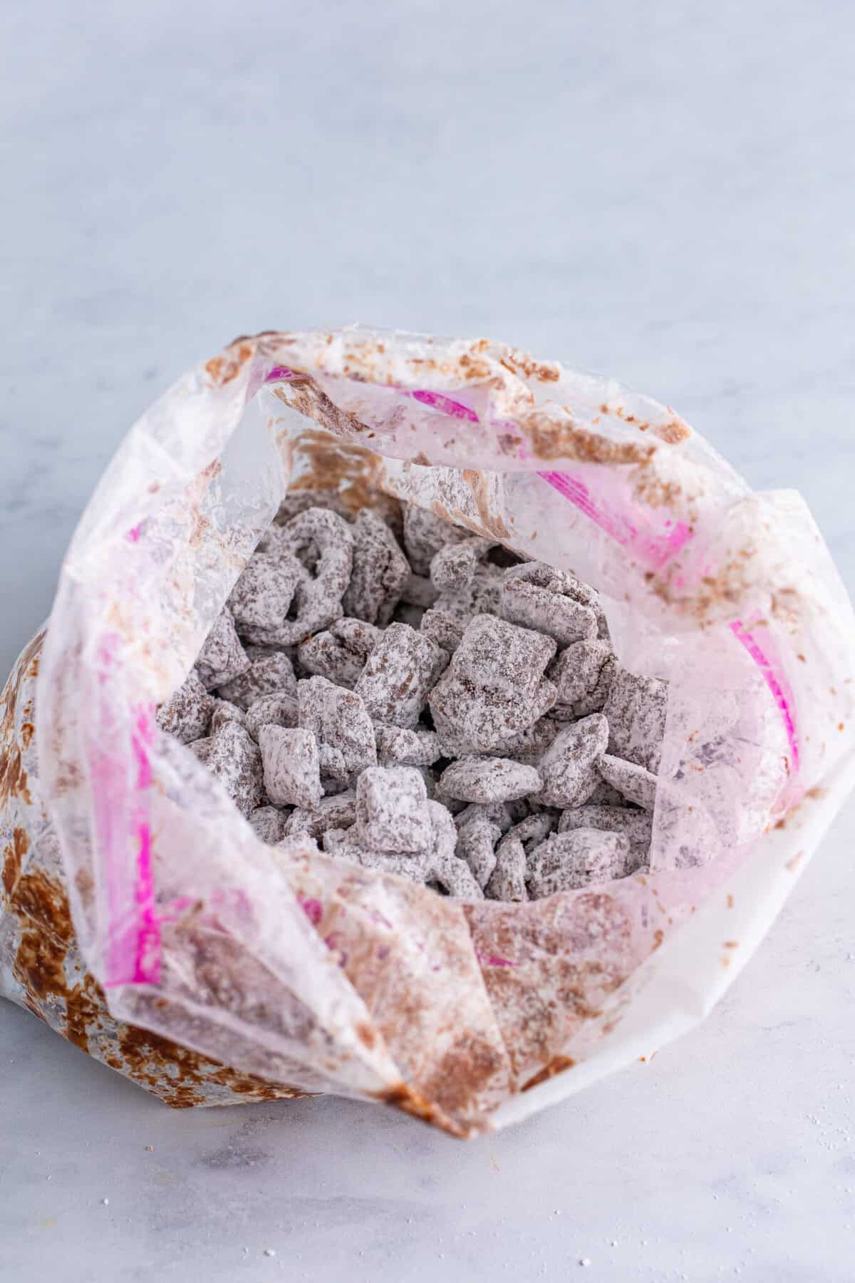 Large pink zip-top bag tossing chocolate-coated Chex cereal with powdered sugar until every piece is dusted white for the Sweet Chex Mix Muddy Buddy.