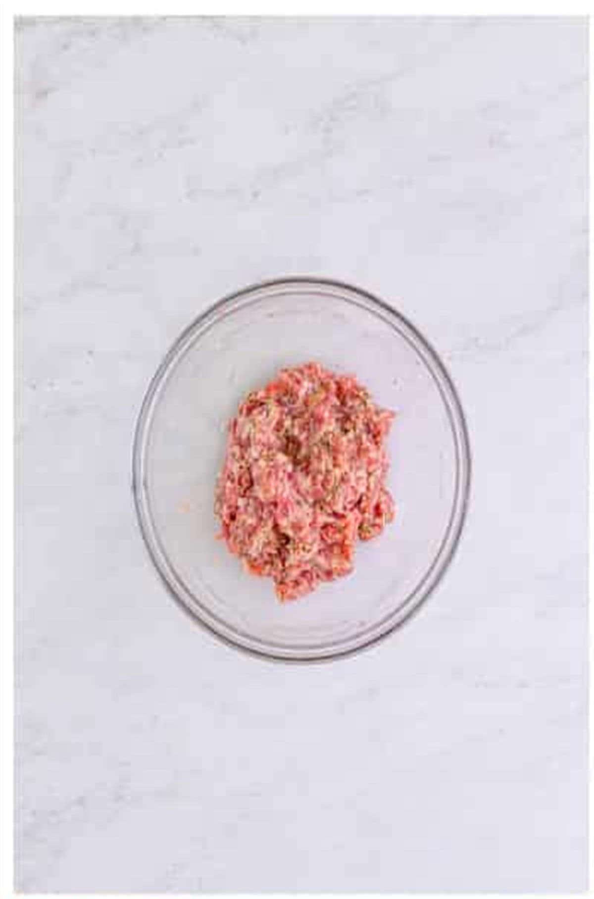 Raw ground pork mixed with crushed fennel, garlic, oregano, basil, salt, and pepper in a glass bowl for The Best Sheet Pan Pizza Recipe.