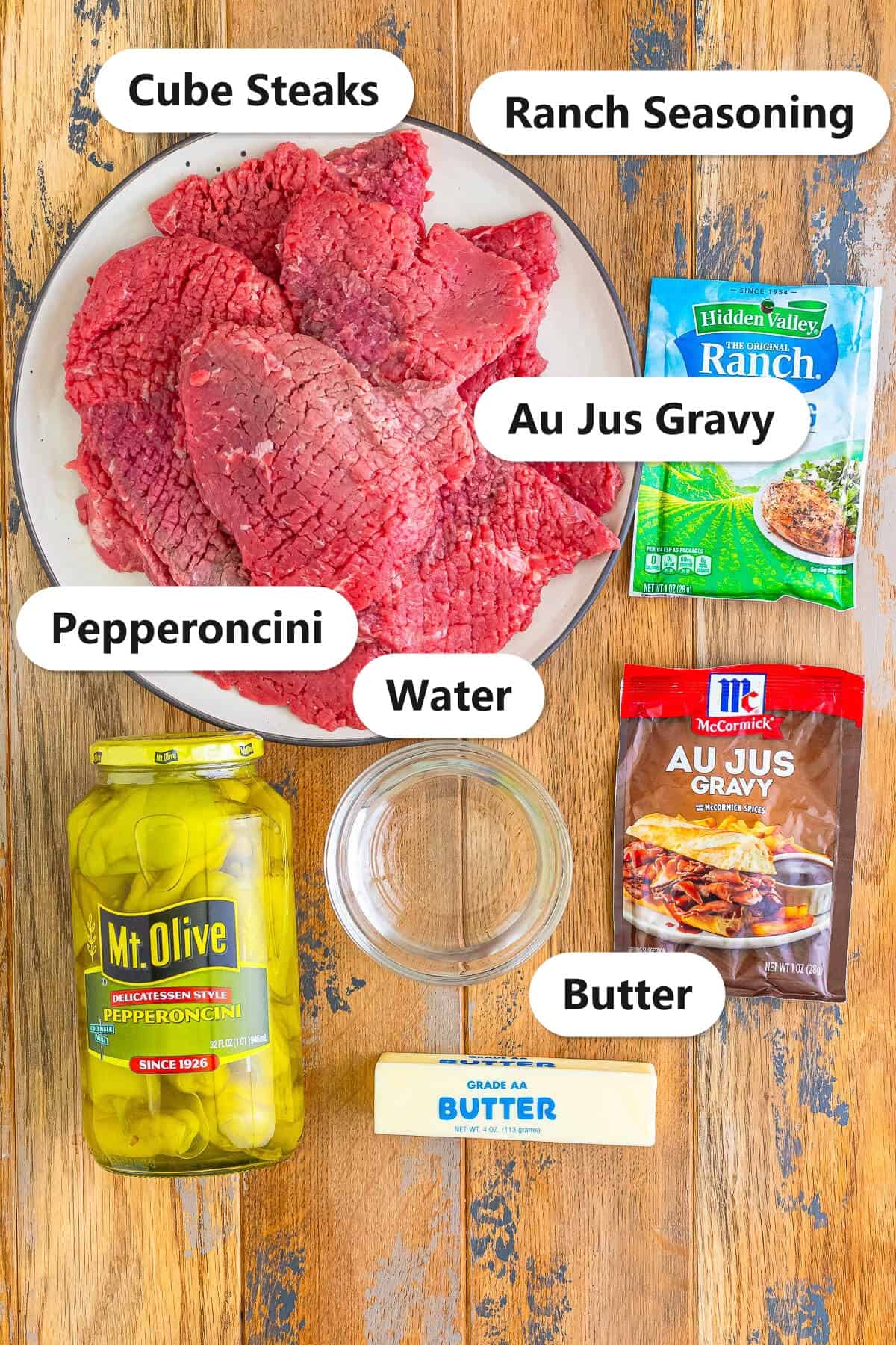 Slow Cooker Cube Steak ingredients labeled on a wooden board including cube steaks, ranch seasoning, au jus gravy mix, pepperoncini, water, and butter.