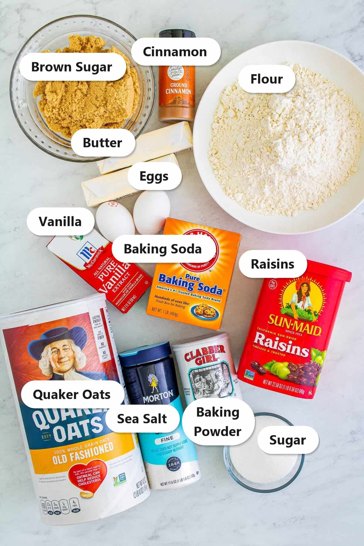 Labeled ingredients flat-lay for Soft Chewy Oatmeal Raisin Cookies (Old Fashioned) including brown sugar, cinnamon, flour, butter, eggs, vanilla, baking soda, raisins, Quaker oats, sea salt, baking powder, and granulated sugar.