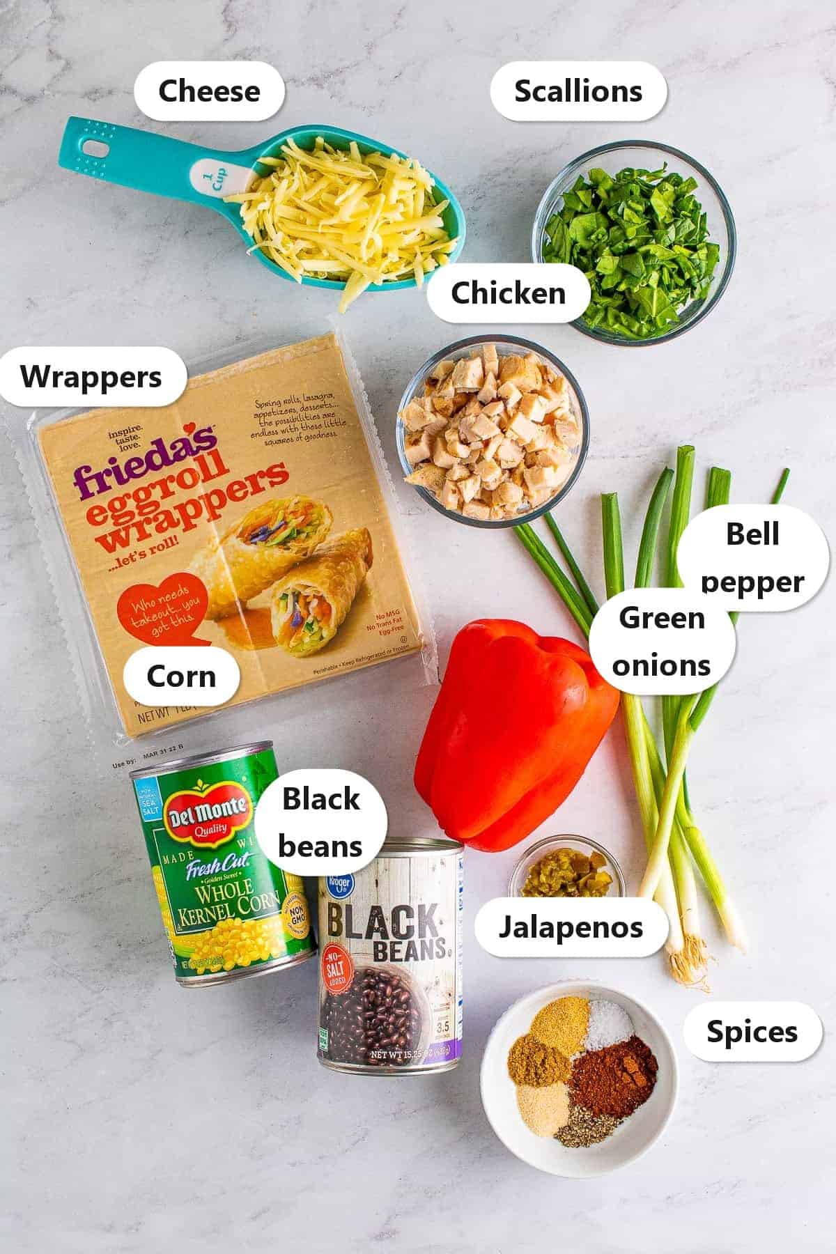 Labeled ingredient flat lay for Copycat Chili Southwest Egg Rolls showing cheese, scallions, Frieda egg roll wrappers, chicken, bell pepper, green onions, corn, black beans, jalapenos, and spices.