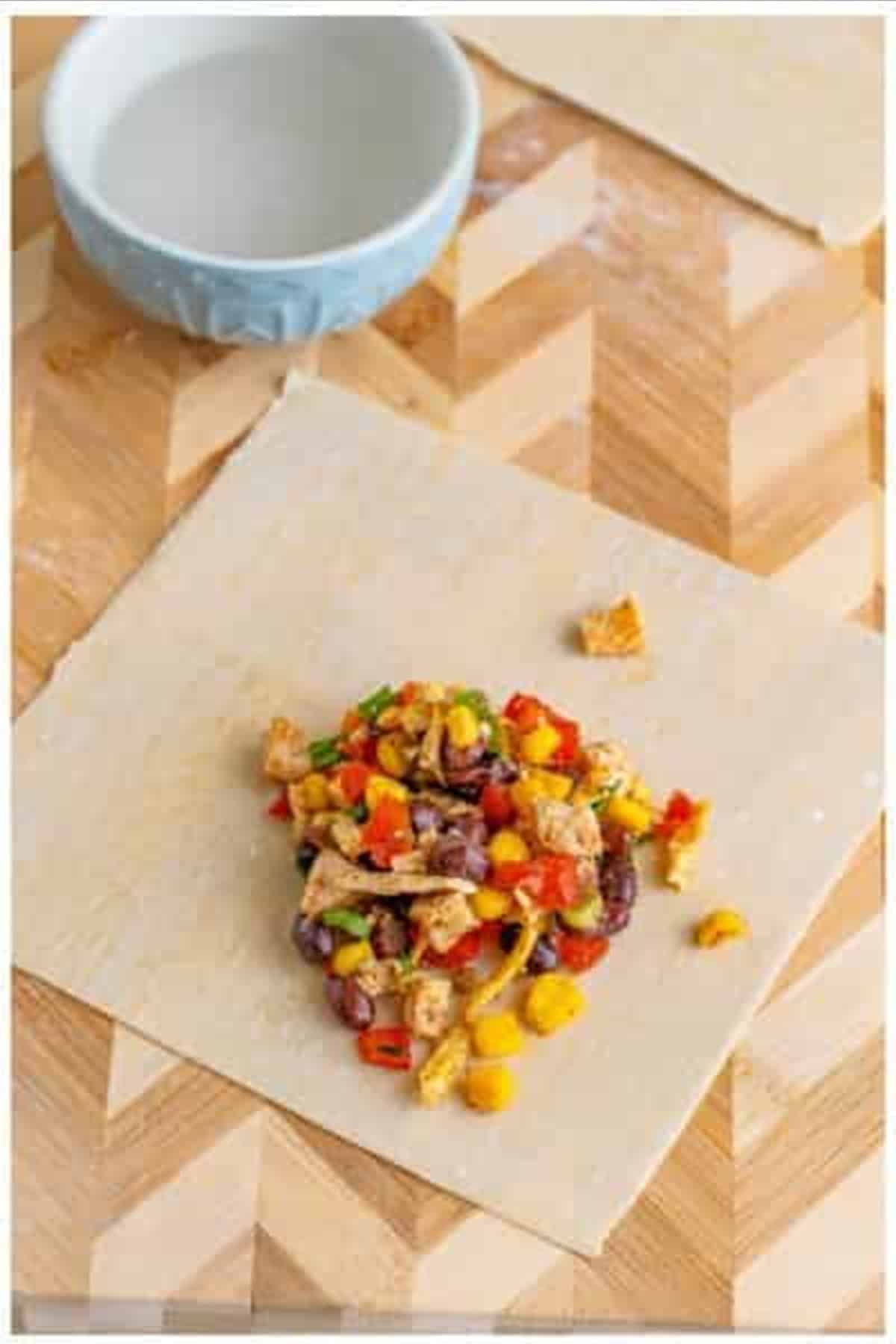 Egg roll wrapper laid out flat with a portion of Southwest filling mixture placed in the center on a wooden cutting board.