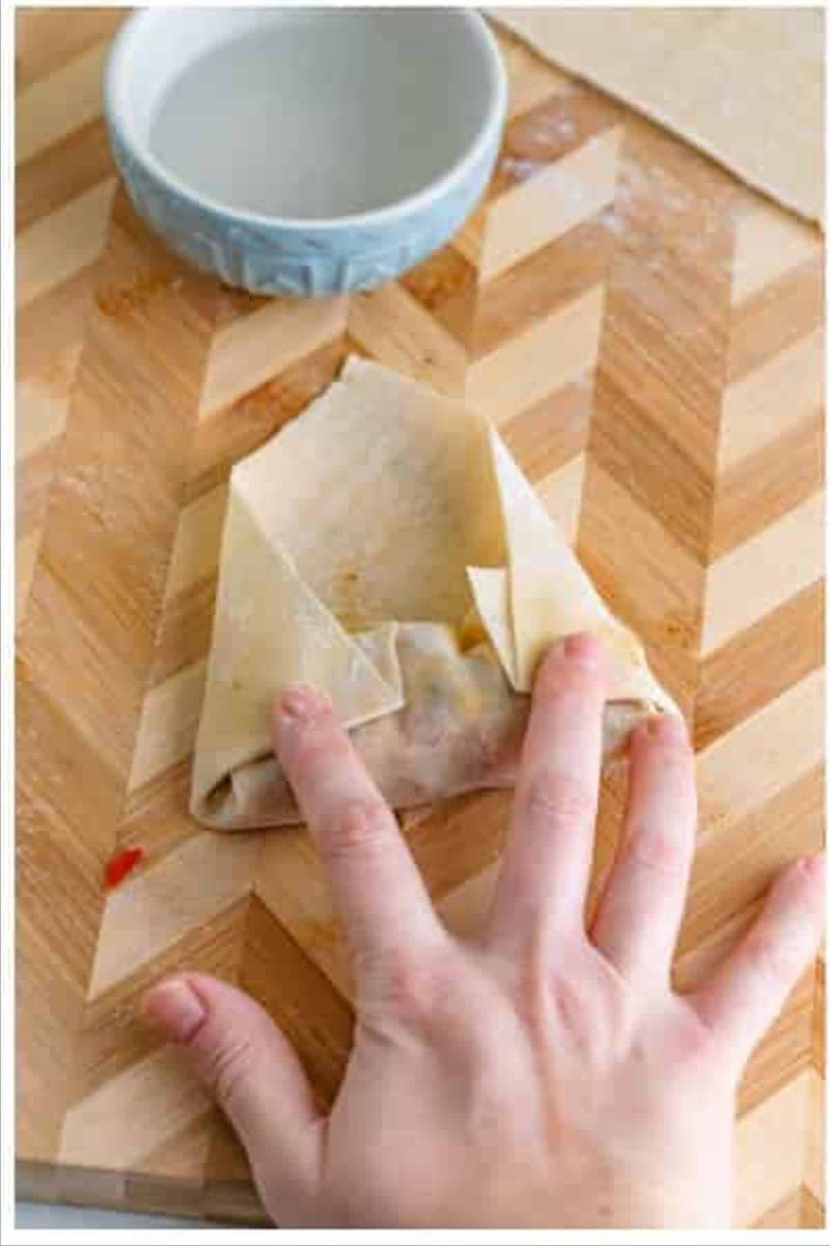 A hand pressing the final edge of a Southwest Egg Roll wrapper sealed with water on a wooden cutting board.