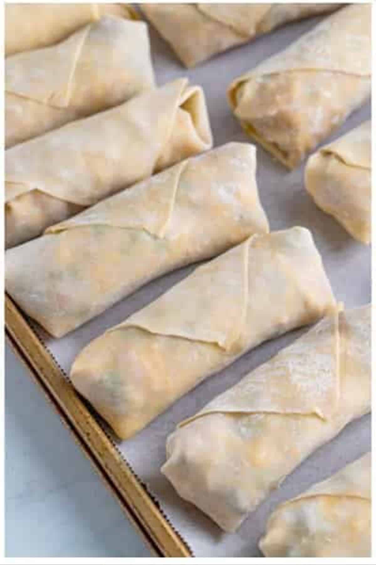Multiple wrapped raw Southwest Egg Rolls arranged in a row on a parchment-lined sheet tray ready to fry.