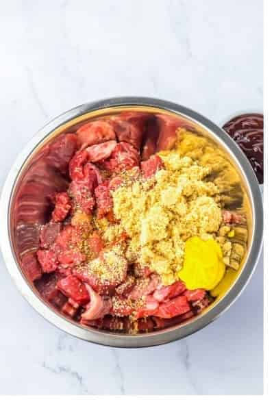 Cubed chuck roast topped with brown sugar, yellow mustard, BBQ rub, and Worcestershire sauce in a metal mixing bowl ready to toss.
