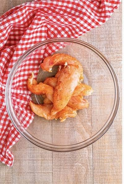 Raw chicken tenders seasoned with Lawry seasoned salt and paprika in a clear glass bowl.