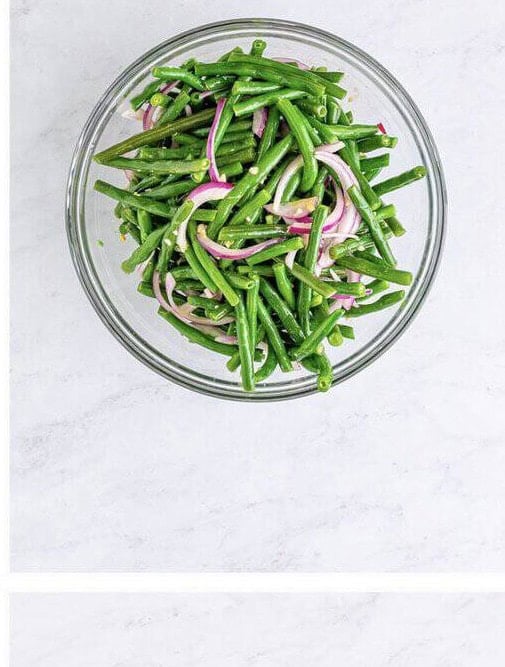 Green beans tossed with red onion and the Dijon vinaigrette in a large mixing bowl, ready for the tomatoes.