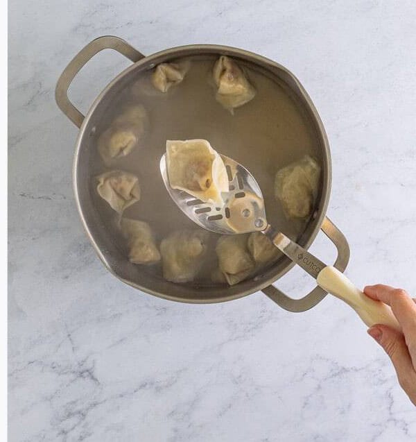 Lifting cooked wontons out of the boiling water with a slotted spoon to transfer to serving bowls.