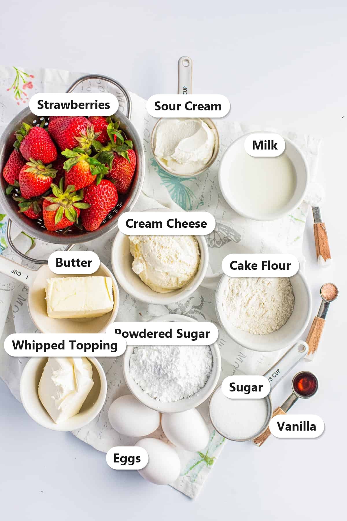 Labeled ingredients for strawberry shortcake cheesecake cake including strawberries, cream cheese, sour cream, milk, butter, cake flour, sugar, powdered sugar, whipped topping, eggs, and vanilla.