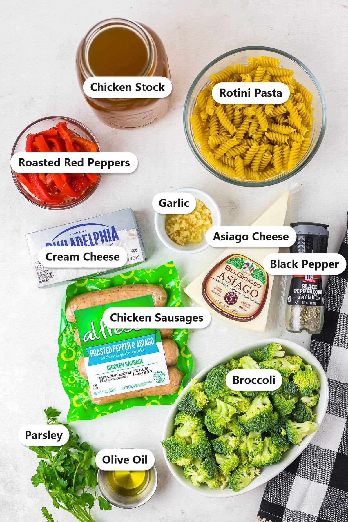 Labeled ingredients for chicken broccoli pasta including rotini, chicken sausages, broccoli, roasted red peppers, asiago, cream cheese, garlic, chicken stock, olive oil, black pepper, and parsley.