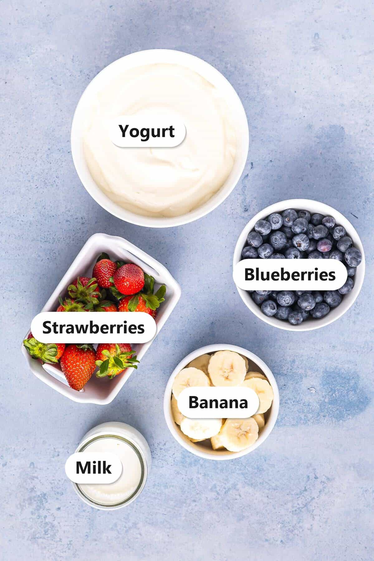 Labeled ingredients for a yogurt fruit smoothie: yogurt, blueberries, strawberries, banana, and milk.
