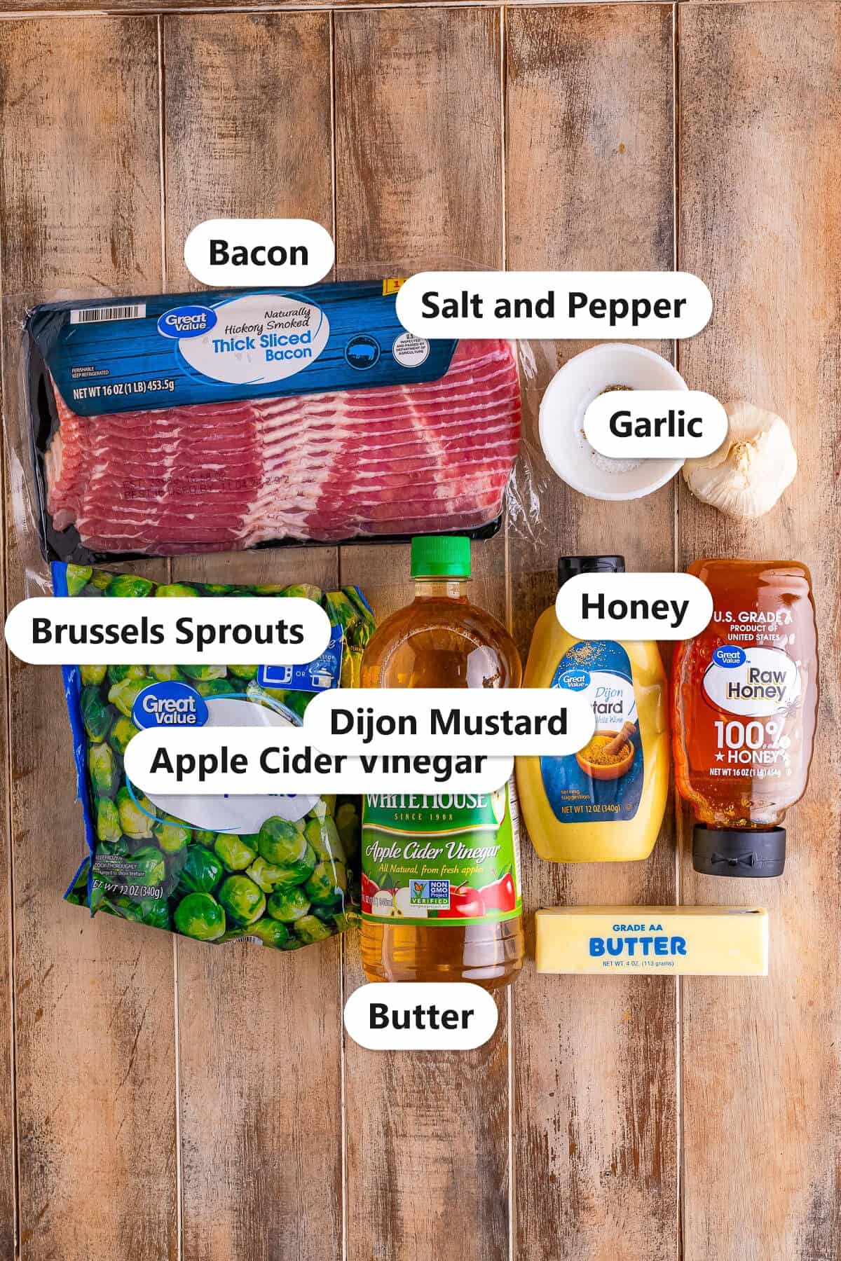 Ingredients to make Brussel Sprouts with Bacon labeled on a wood surface including bacon, frozen Brussels sprouts, apple cider vinegar, dijon mustard, honey, butter, salt, pepper, and garlic.