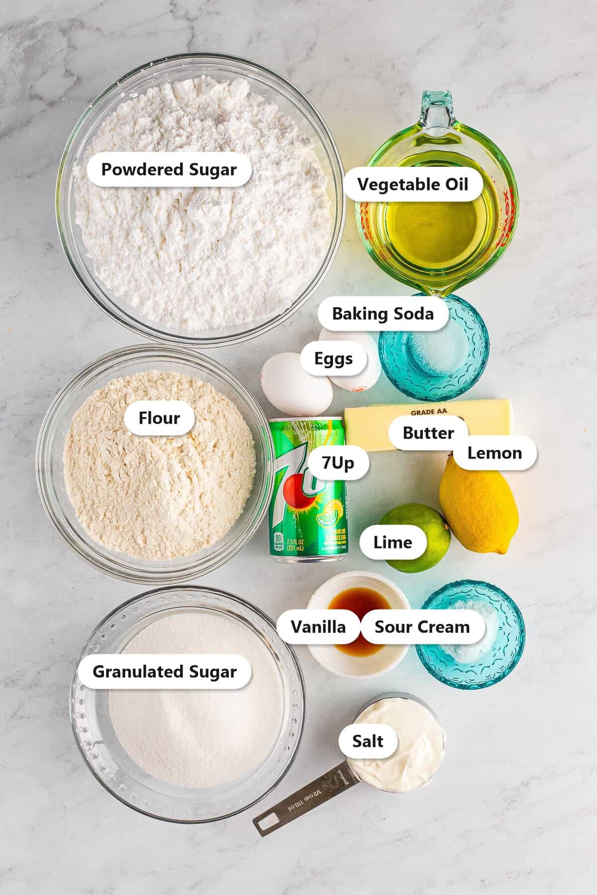 Labeled ingredients for 7Up cake including flour, granulated sugar, powdered sugar, 7Up, vegetable oil, sour cream, eggs, butter, lemon, lime, and vanilla.