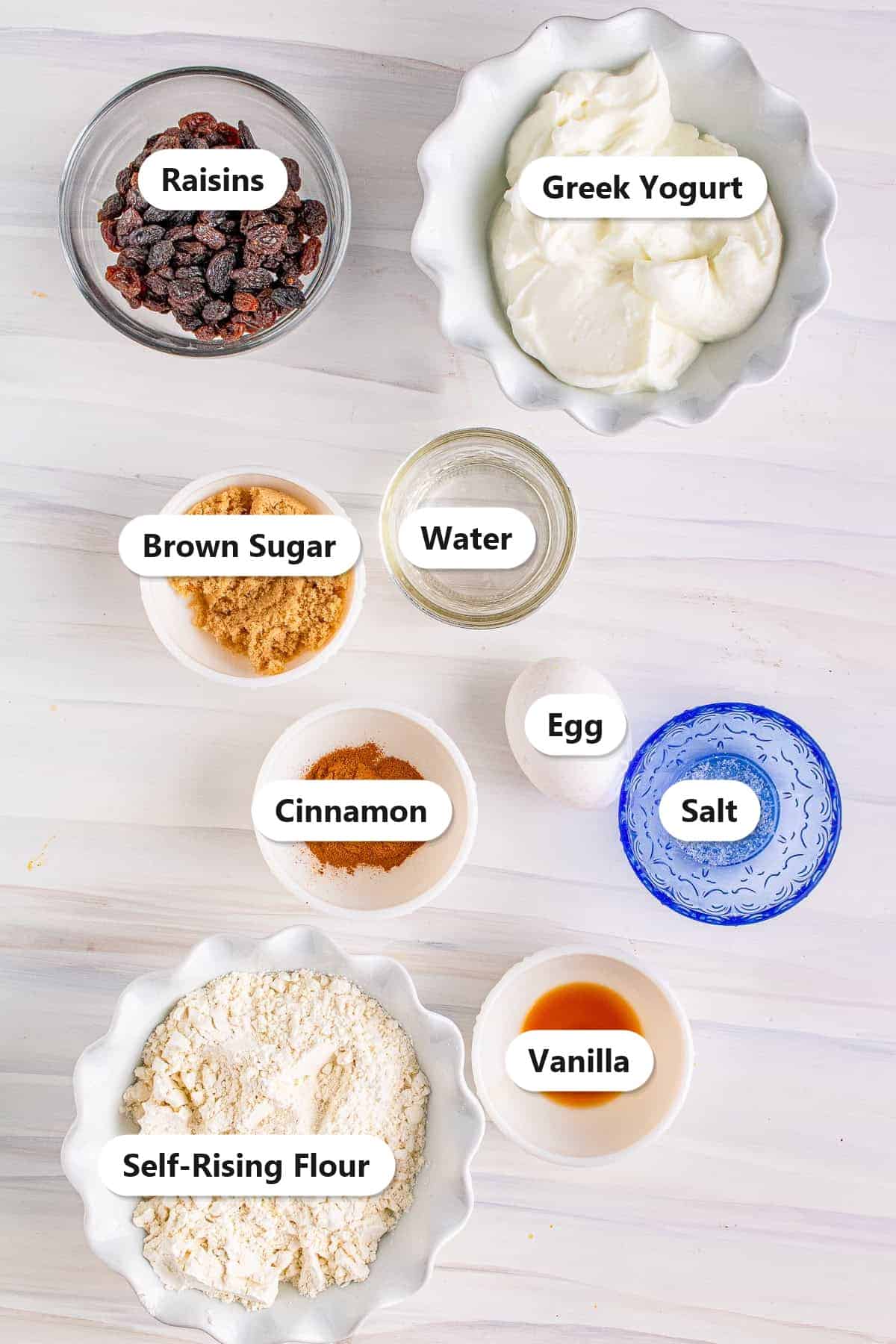 Labeled ingredients for cinnamon raisin air fryer bagels including self-rising flour, Greek yogurt, raisins, brown sugar, cinnamon, and egg.