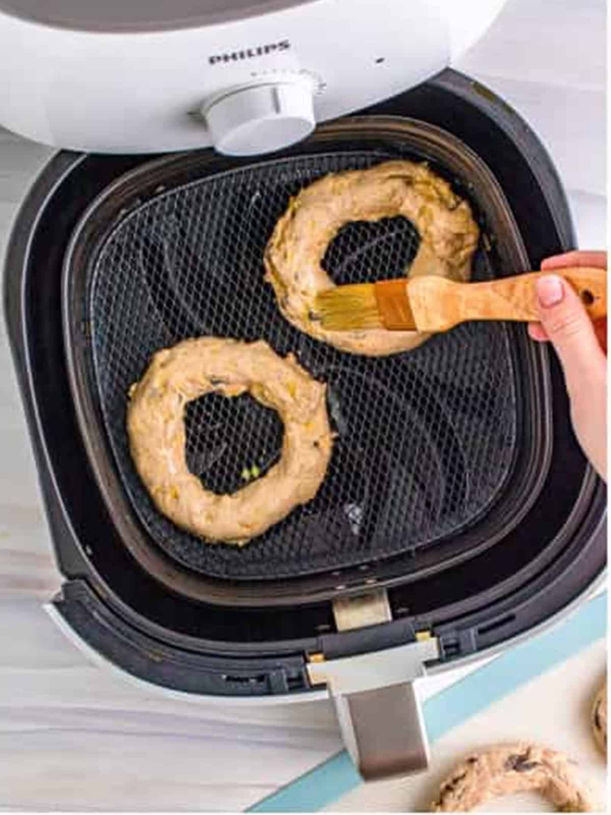 Brushing egg wash over cinnamon raisin bagels in the air fryer basket.