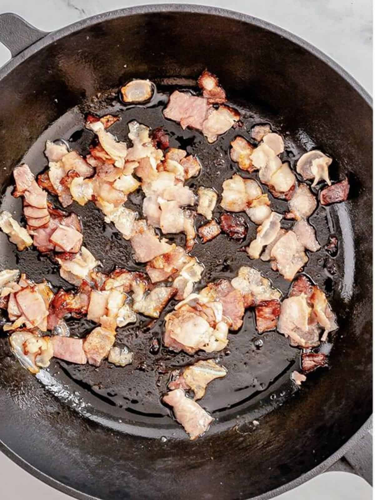 Diced bacon cooking until crispy in a cast iron skillet.