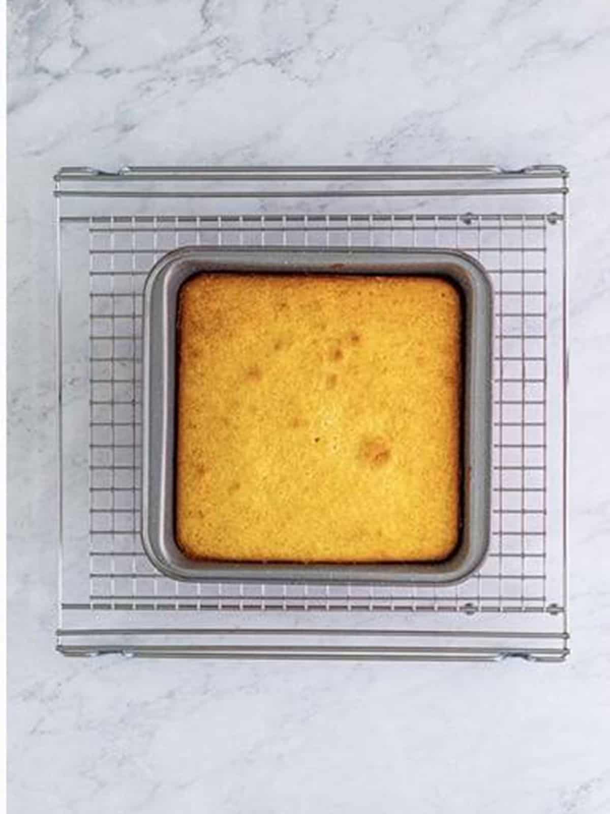 A golden baked almond cake cooling in a square pan on a wire rack.