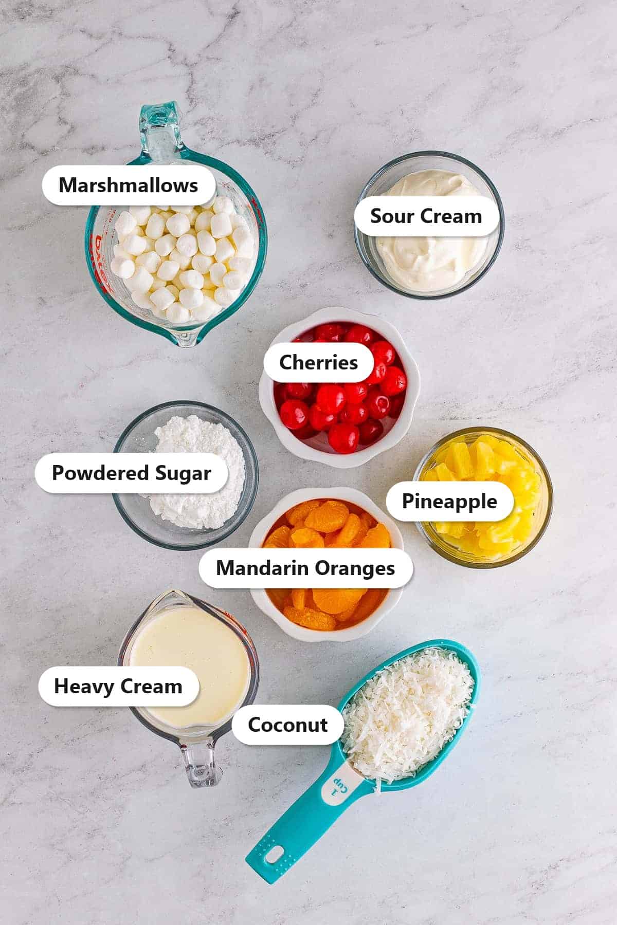 Ambrosia salad ingredients including marshmallows, sour cream, cherries, pineapple, mandarin oranges, coconut, heavy cream, and powdered sugar labeled on a marble surface.