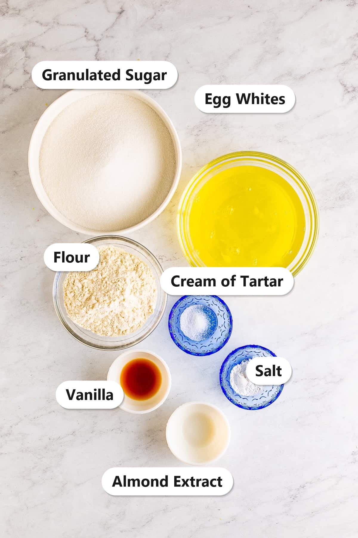 Labeled angel food cake ingredients including sugar, flour, egg whites, cream of tartar, salt, vanilla, and almond extract on a marble counter.