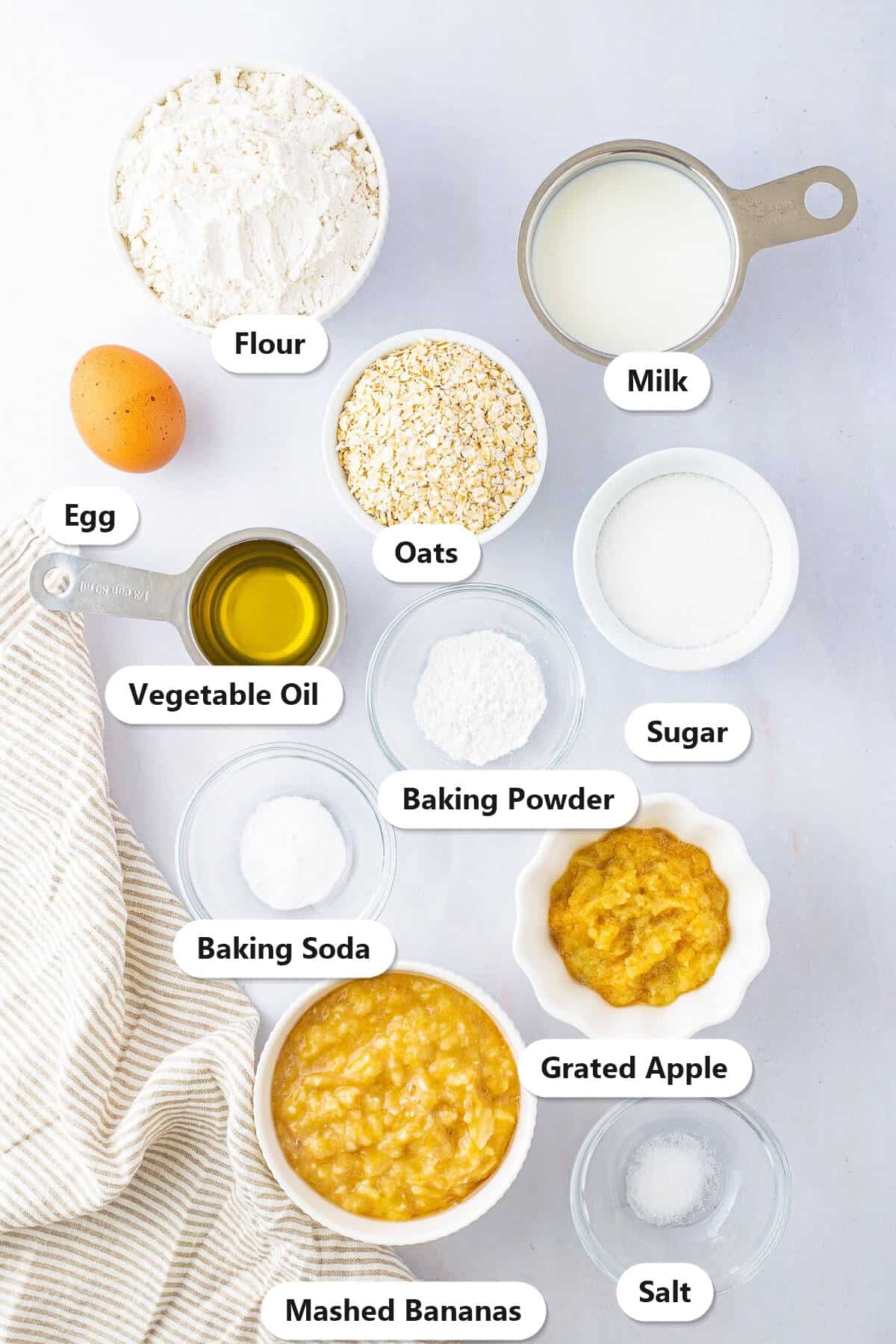 Labeled ingredients for apple banana muffins including flour, oats, sugar, milk, egg, vegetable oil, mashed bananas, grated apple, baking powder, baking soda, and salt.