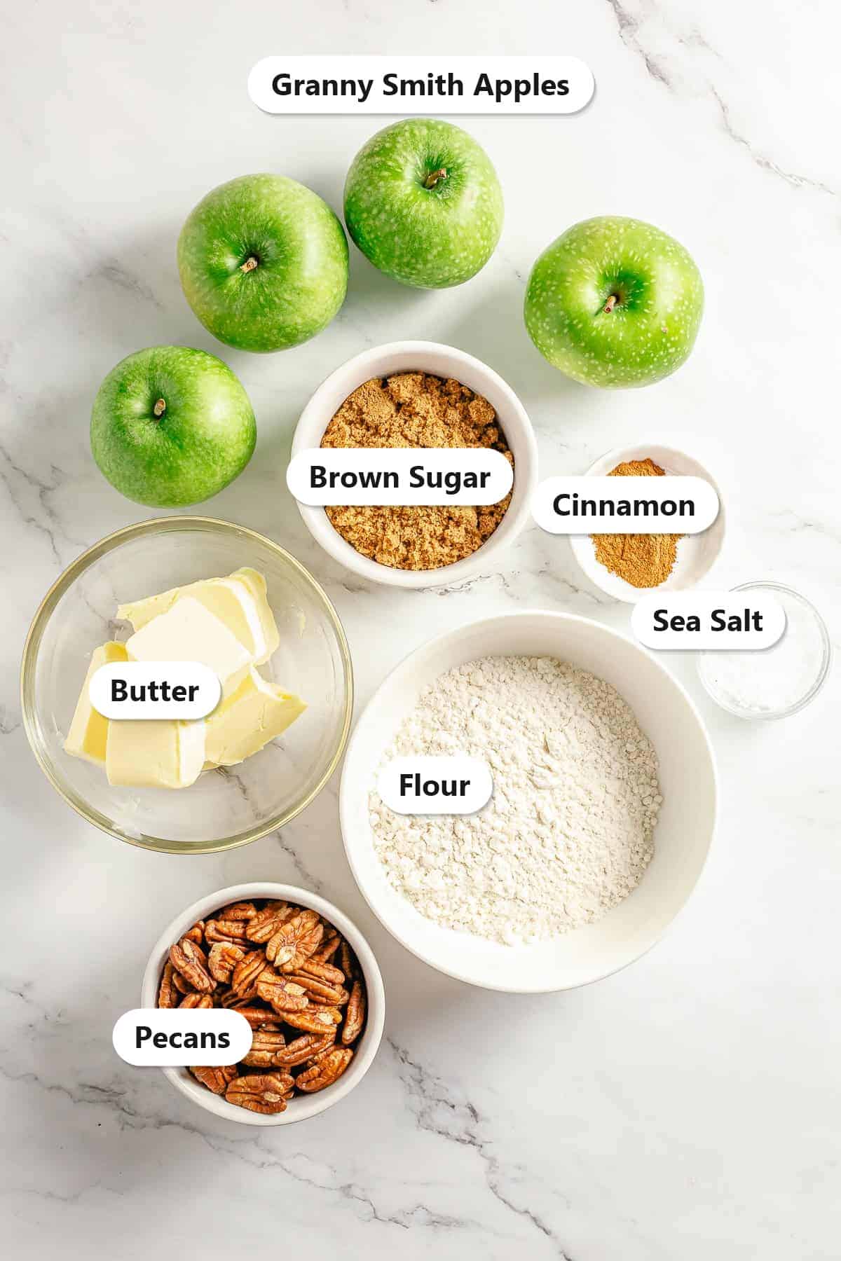 Labeled ingredients to make apple crumble: granny smith apples, brown sugar, cinnamon, sea salt, butter, flour, and pecans.