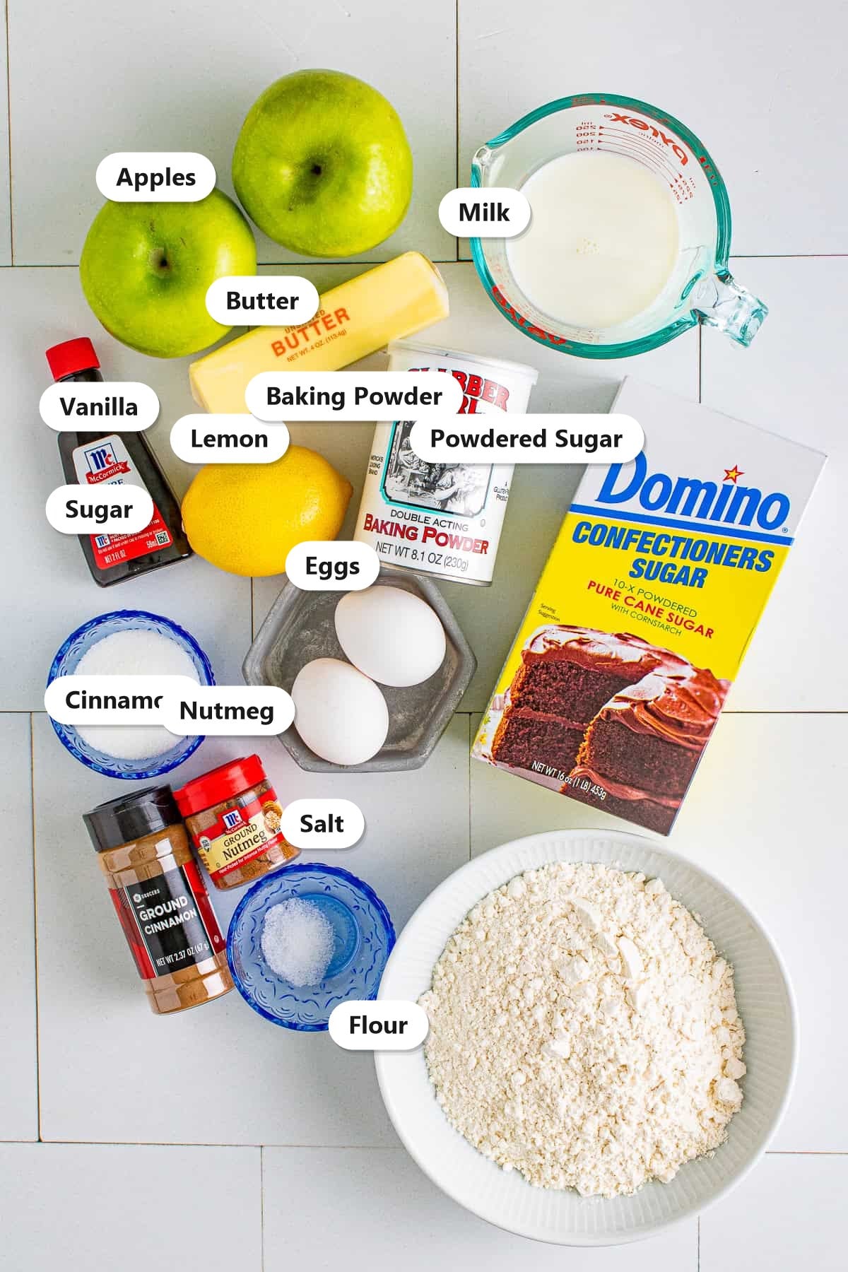 Apple fritter ingredients including Granny Smith apples, flour, milk, butter, eggs, sugar, cinnamon, nutmeg, vanilla, and powdered sugar labeled on a tile surface.