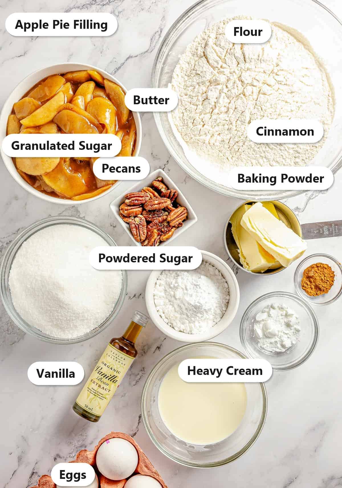 Labeled ingredients for apple pie bars including apple pie filling, flour, butter, granulated sugar, powdered sugar, eggs, pecans, cinnamon, vanilla, baking powder, and heavy cream.